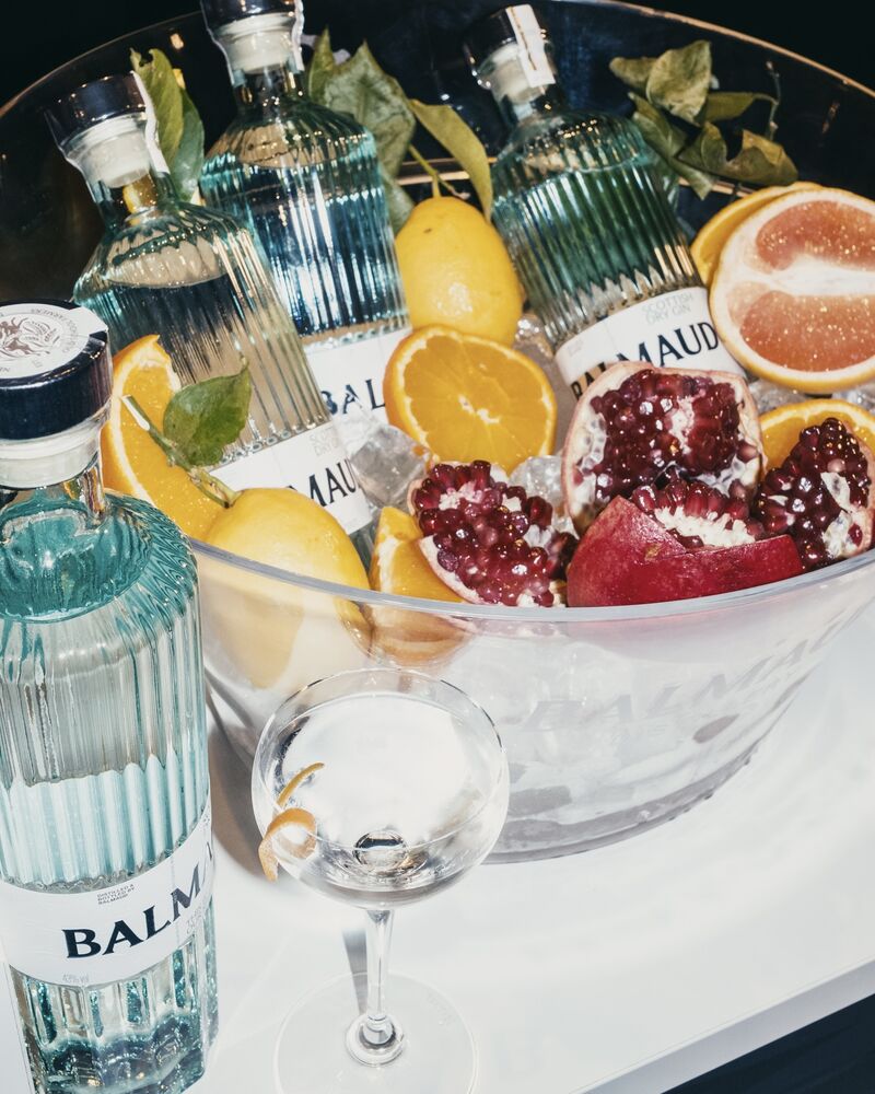 A styled cocktail setup featuring Balma and Maudoux bottles, a large glass ice bucket filled with citrus slices, pomegranate halves, and green leaves, alongside an empty coupe glass on a white surface.