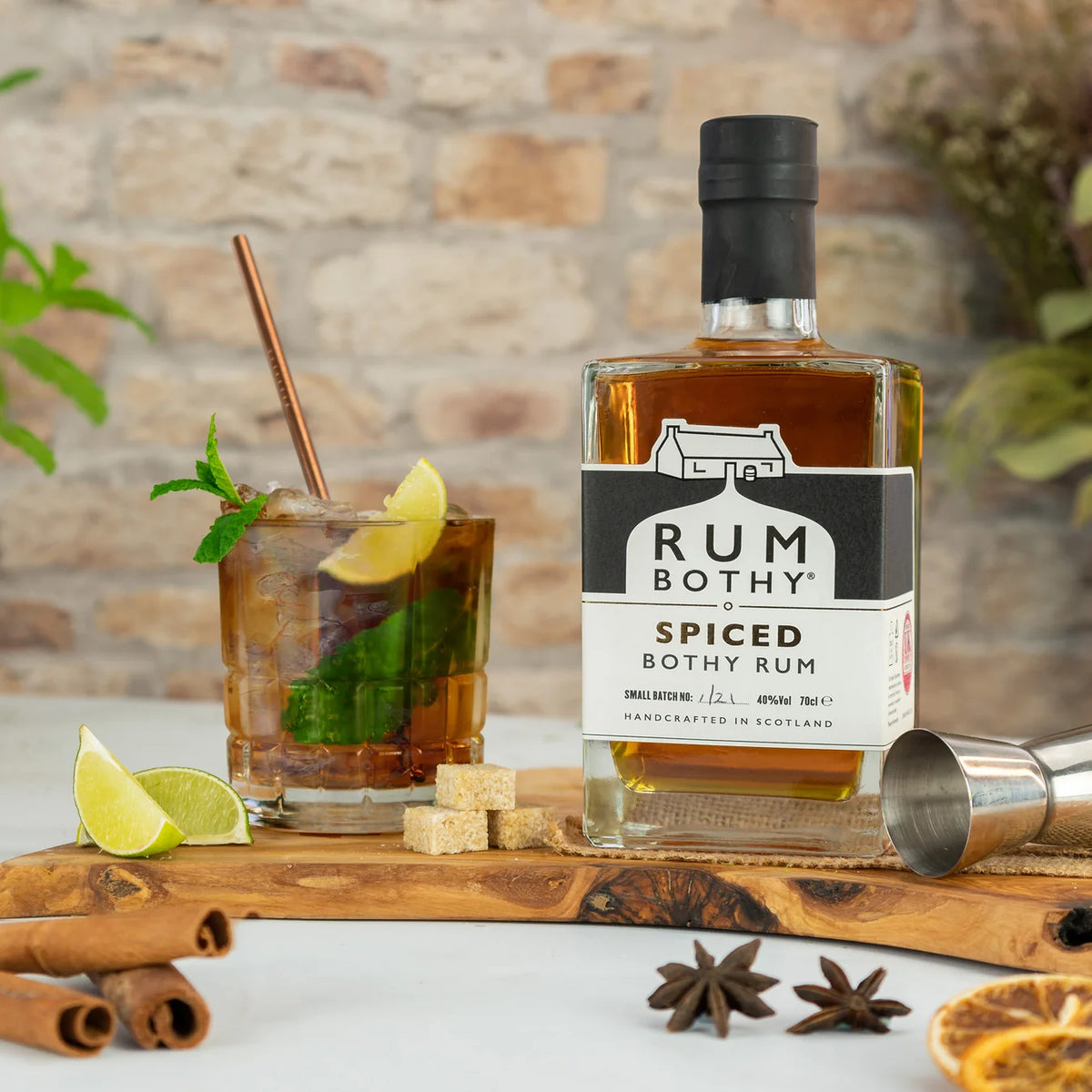 A bottle of Rum Bothy Spiced Bothy Rum stands on a wooden board beside a prepared cocktail glass with lime, a metal jigger, cinnamon sticks, and star anise, set against a blurred brick wall background.